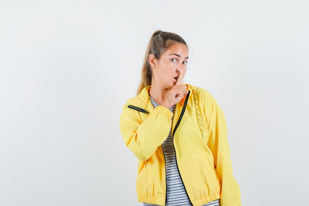 Photo gratuite femme blonde montrant le geste de silence en blouson aviateur jaune et chemise rayée et à la recherche concentrée