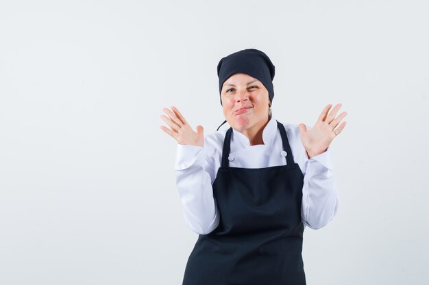 Femme blonde montrant un panneau d'arrêt à deux mains, grimaçant en uniforme de cuisinier noir et à la jolie. vue de face.