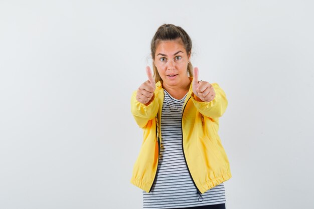 Femme blonde montrant les pouces vers le haut avec les deux mains en veste bomber jaune et chemise rayée et à la jolie