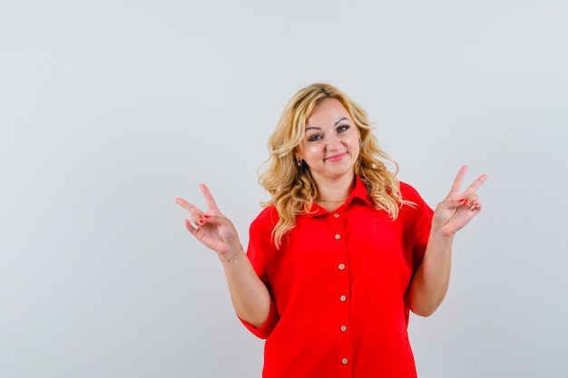 Femme blonde montrant le signe de la paix avec les deux mains en chemisier rouge et à la recherche de bonheur.