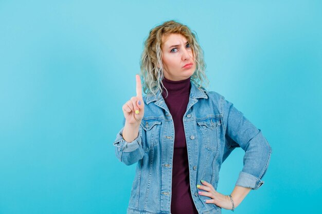 Une femme blonde montre un geste minute et met l'autre main sur la taille sur fond bleu
