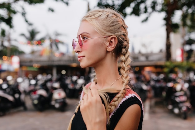 Femme blonde pensive en tenue noire posant sur fond de rue flou. dame bronzée avec des tresses porte des lunettes de soleil roses.