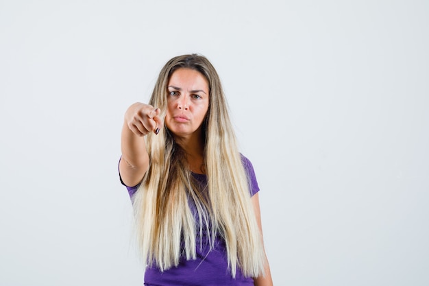 femme blonde pointant vers l'avant en t-shirt violet et à la recherche de sérieux. vue de face.