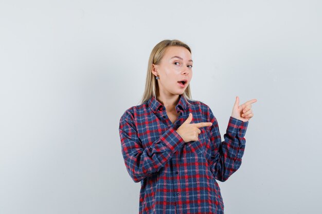 Femme blonde pointant vers la droite avec l'index en chemise à carreaux et à l'optimiste, vue de face.