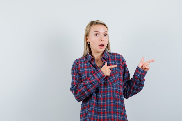 Femme blonde pointant vers la droite avec l'index en chemise à carreaux et à la surprise, vue de face.