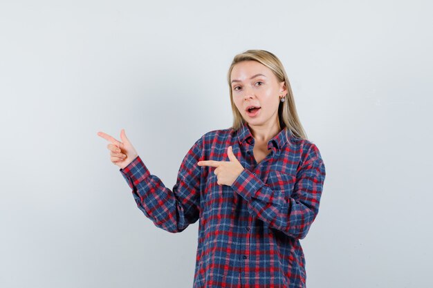 Femme blonde pointant vers la gauche avec l'index en chemise à carreaux et à la surprise, vue de face.