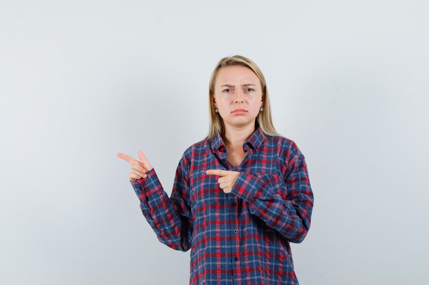 Femme blonde pointant vers la gauche avec l'index et grimaçant en chemise à carreaux et à la mécontentement. vue de face.