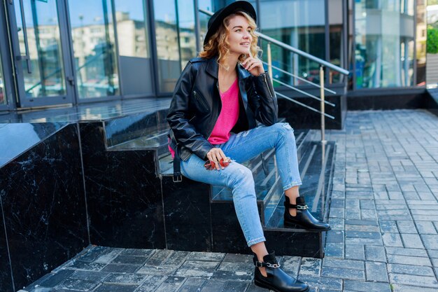 femme blonde posant dans les rues modernes. Tenue d'automne élégante, veste en cuir et pull tricoté. Lunettes de soleil roses.