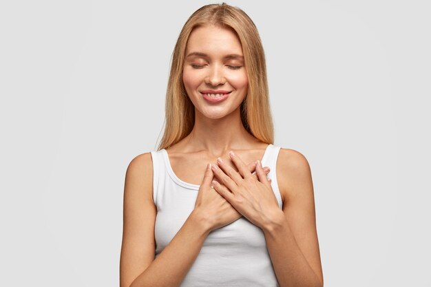 Femme blonde positive optimiste avec les mains sur la poitrine, vêtue d'un gilet décontracté, a un sourire charmant