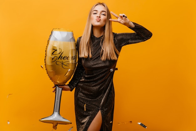 Femme blonde positive posant avec un grand verre à vin. Adorable femme blanche en robe préparant la fête d'anniversaire.