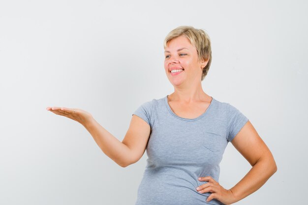 Femme blonde qui s'étend des mains comme présentant quelque chose en t-shirt bleu clair et à la gaieté. vue de face.