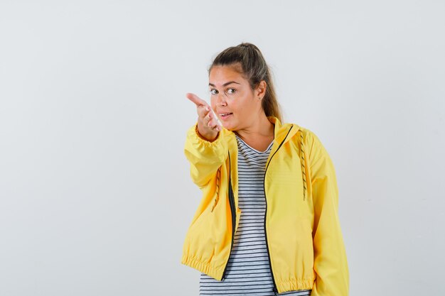 Femme blonde qui tend la main vers l'avant comme invitant à venir en blouson aviateur jaune et chemise rayée et à la jolie