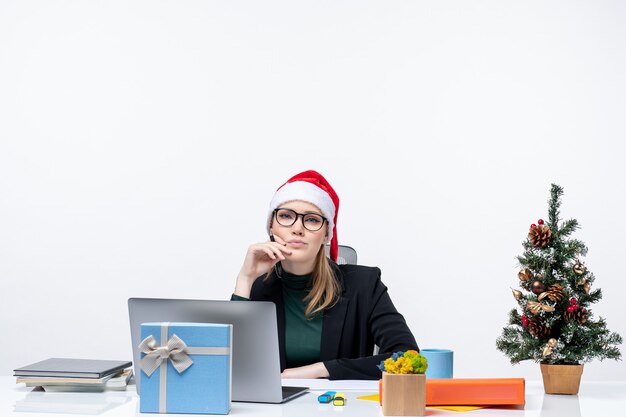 Femme blonde réfléchie avec un chapeau de père Noël assis à une table avec un arbre de Noël et un cadeau dessus dans le bureau sur fond blanc