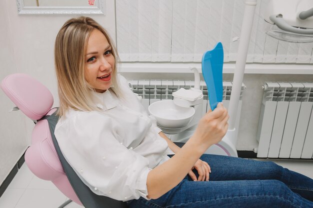 Une femme blonde regarde ses dents dans le miroir, assise sur une chaise. jeune fille souriante au bureau du dentiste