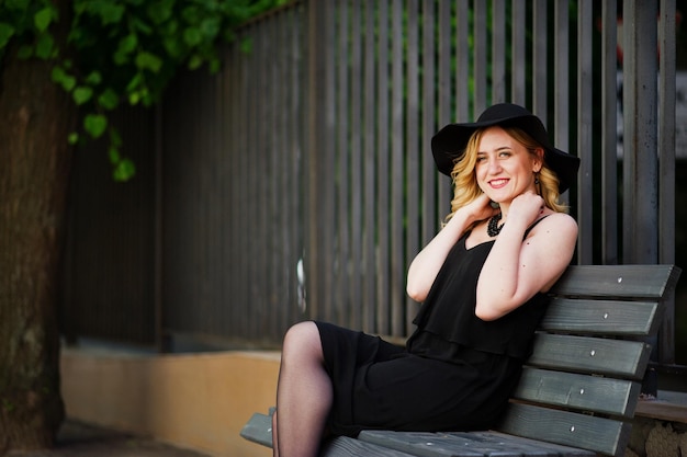 Femme blonde sur robe noire colliers et chapeau assis sur un banc