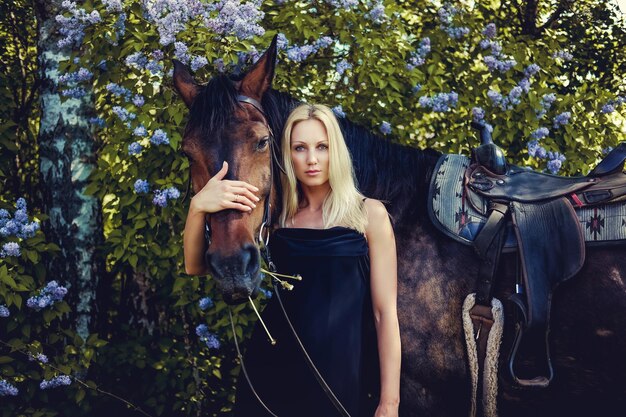 Femme blonde en robe de soirée noire posant avec un cheval brun.