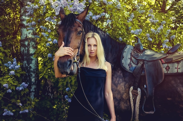 Femme blonde en robe de soirée noire posant avec un cheval brun.
