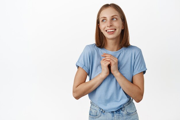 Femme blonde romantique rêvant de qch, se tenant la main près du cœur, pensant à quelqu'un qu'elle aime, souriant rêveuse et regardant le logo du coin supérieur gauche, mur blanc.