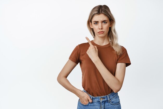 Femme blonde sceptique pointant le doigt dans le coin supérieur gauche et fronçant les sourcils contrarié montrant qch peu amusant debout en t-shirt sur fond blanc