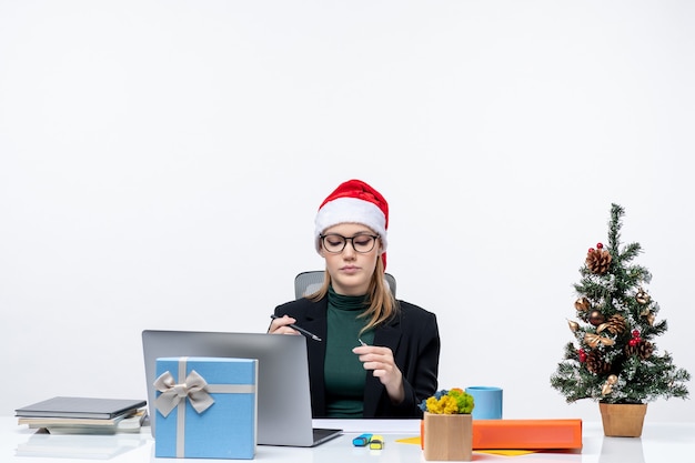 Femme blonde sérieuse avec un chapeau de père Noël assis à une table avec un arbre de Noël et un cadeau dessus dans le bureau sur fond blanc