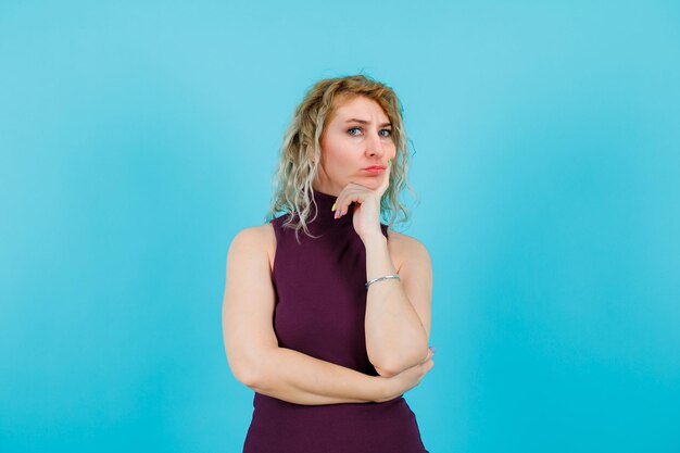 Une femme blonde sérieuse regarde la caméra en mettant la main sur le menton sur fond bleu