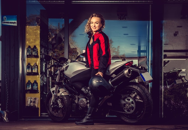 Femme blonde sexy en veste de cuir rouge posant près de la moto.