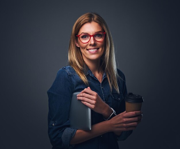 Une femme blonde souriante à lunettes tient une tablette PC et emporte une tasse de café.