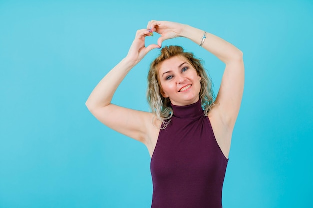 Une femme blonde souriante montre un geste cardiaque en se tenant la main au-dessus de la tête sur fond bleu