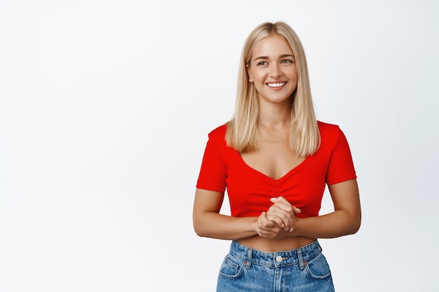 Femme blonde souriante tenant les mains ensemble regardant de côté avec une expression de visage agréable et amicale fond blanc portant un haut court et un jean