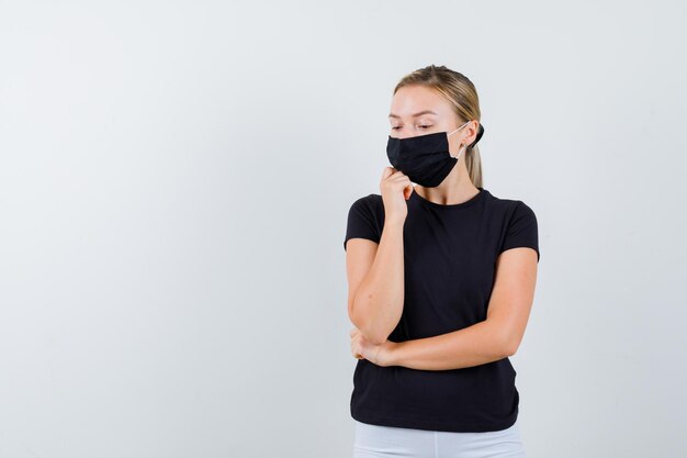 Femme blonde soutenant le menton sur le poing, debout dans une pose de réflexion en t-shirt noir