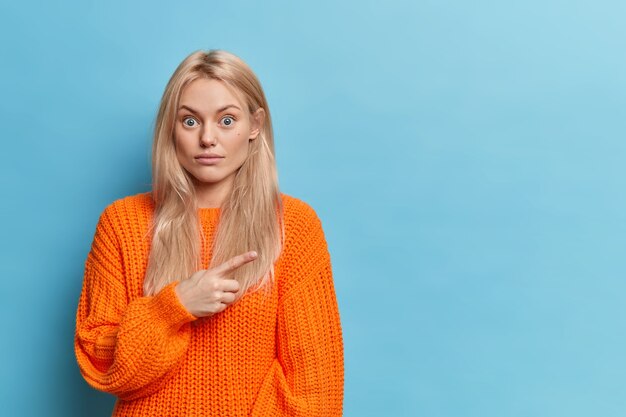 Femme blonde surprise pointe à droite sur l'espace de copie sur le mur bleu, exprime la surprise