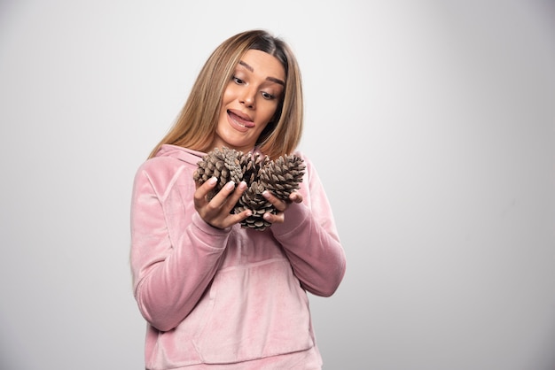 Une femme blonde en sweat-shirt rose fait des visages positifs idiots avec des cônes de chêne dans la main.