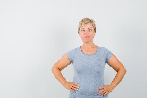 Femme blonde en t-shirt bleu clair tenant ses mains sur la taille et à la vue de face, confiant.