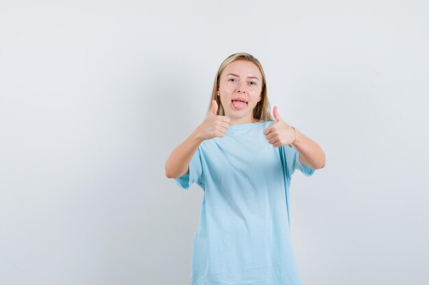 Femme blonde en t-shirt bleu montrant le double pouce vers le haut, tirant la langue