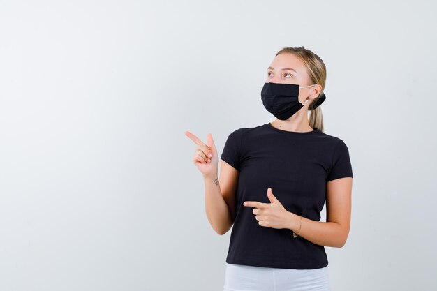 Femme blonde en t-shirt noir, pantalon blanc, masque noir pointant vers la gauche avec l'index