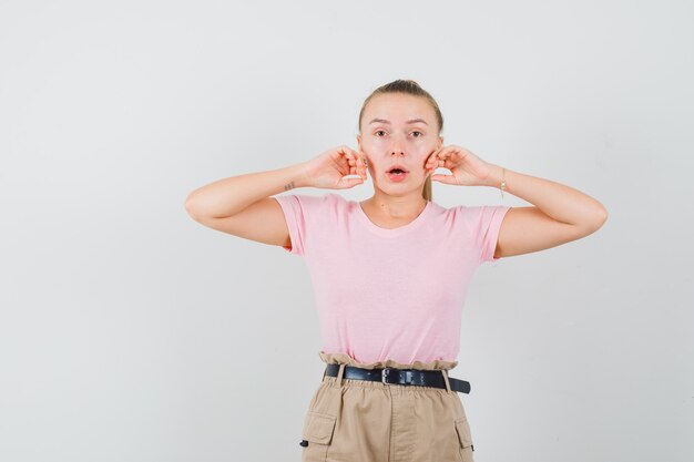 Femme blonde en t-shirt, pantalon touchant les joues avec les mains et à la surprise, vue de face.