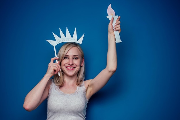 Femme blonde tenant un drapeau américain avec une couronne de papier et une torche statue de la liberté sur un fond bleu dans le studio. concept de célébration de la fête de l'indépendance du 4 juillet et apprentissage de la langue anglaise