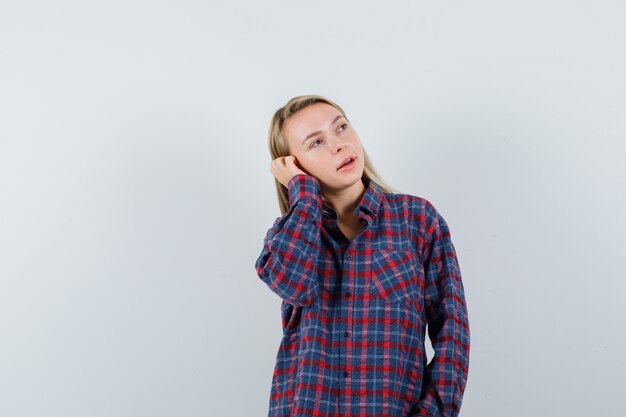 Femme blonde tenant la main sur l'oreille, faisant semblant de parler au téléphone en chemise à carreaux et à la recherche concentrée. vue de face.