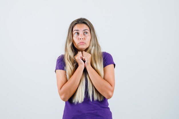 femme blonde tenant les poings ensemble sur la poitrine en t-shirt violet et à la peur, vue de face.