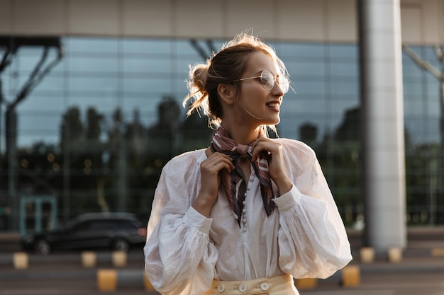 Photo gratuite femme blonde touche son foulard marron en soie jolie jeune fille à lunettes et chemisier blanc sourit à l'extérieur