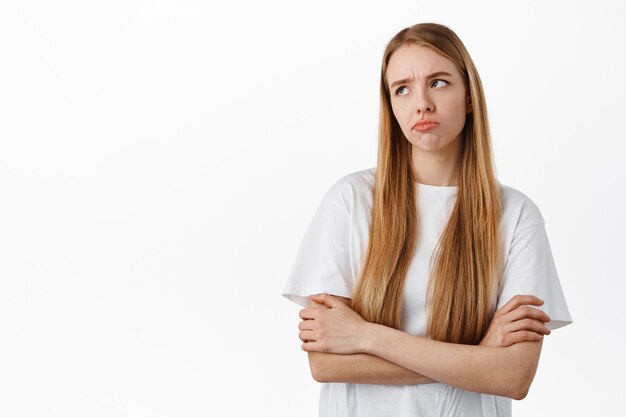 Femme blonde triste et sombre boudant, grimaçant mécontent, les bras croisés sur la poitrine réticents et regardant le logo du coin supérieur gauche, debout sur un mur blanc