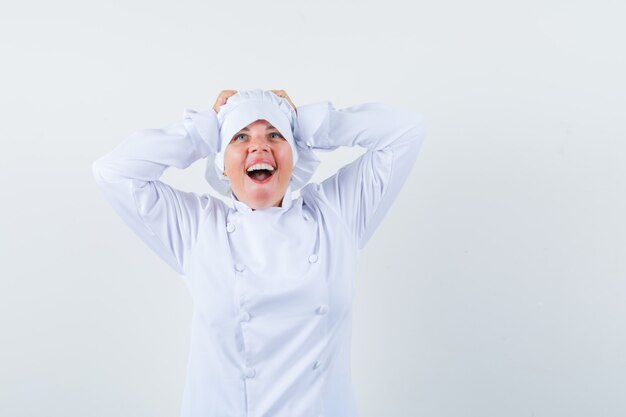 Femme blonde en uniforme de cuisinier blanc tenant les mains sur la tête, se réjouissant et à la jolie