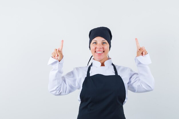 Femme blonde en uniforme de cuisinier noir pointant vers le haut avec l'index et à la jolie