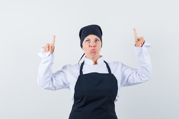 Femme blonde en uniforme de cuisinier noir pointant vers le haut avec l'index et à la jolie