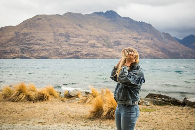 Femme blonde avec une veste en jean debout près de la mer ondulée entourée de hautes montagnes rocheuses