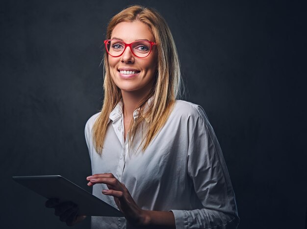 Une femme blonde vêtue d'une chemise blanche et de lunettes tient une tablette PC sur fond gris.