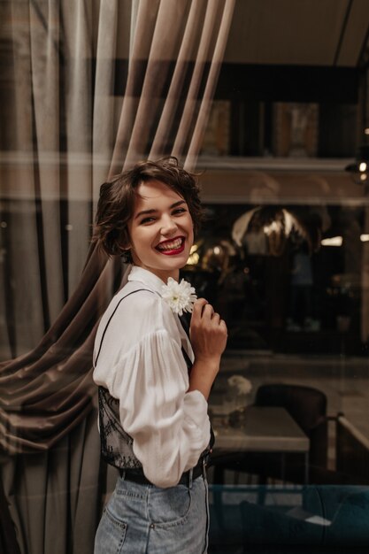 Femme de bonne humeur aux cheveux brune en chemise blanche riant à l'intérieur. Dame aux cheveux courts en jeans avec des lèvres rouges tenant une fleur au café.