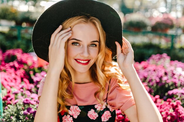 Femme de bonne humeur aux grands yeux posant dans le jardin. Jocund femme au chapeau debout en frint de fleurs roses.