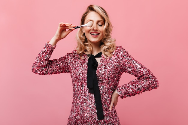 Femme de bonne humeur posant avec un pinceau de maquillage sur un mur rose