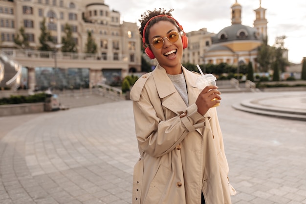 Une femme de bonne humeur en trench-coat beige surdimensionné boit du jus d'orange, sourit et écoute de la musique dans des écouteurs rouges à l'extérieur